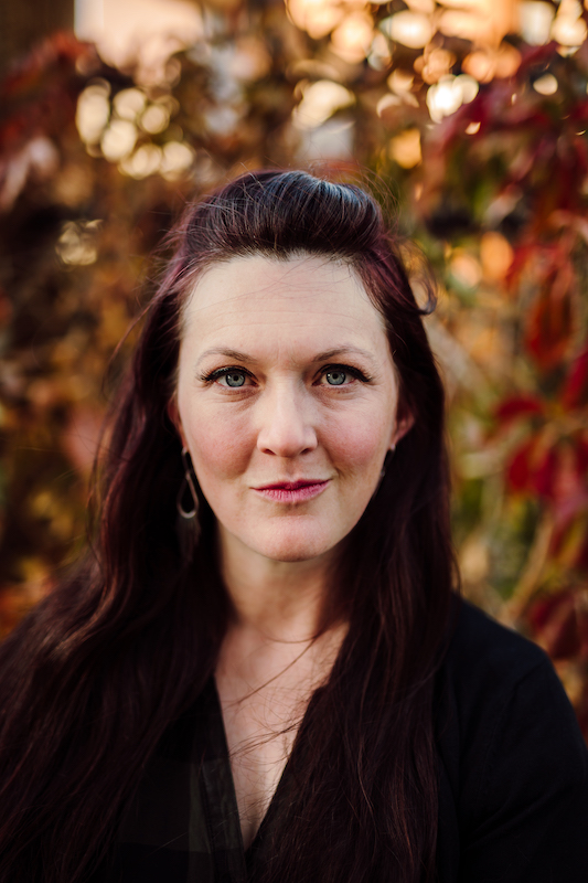 Image is a photo of writer Crystal J. Bell, a white woman with long dark hair wearing a dark top, with blurry autumnal-looking foliage in the background