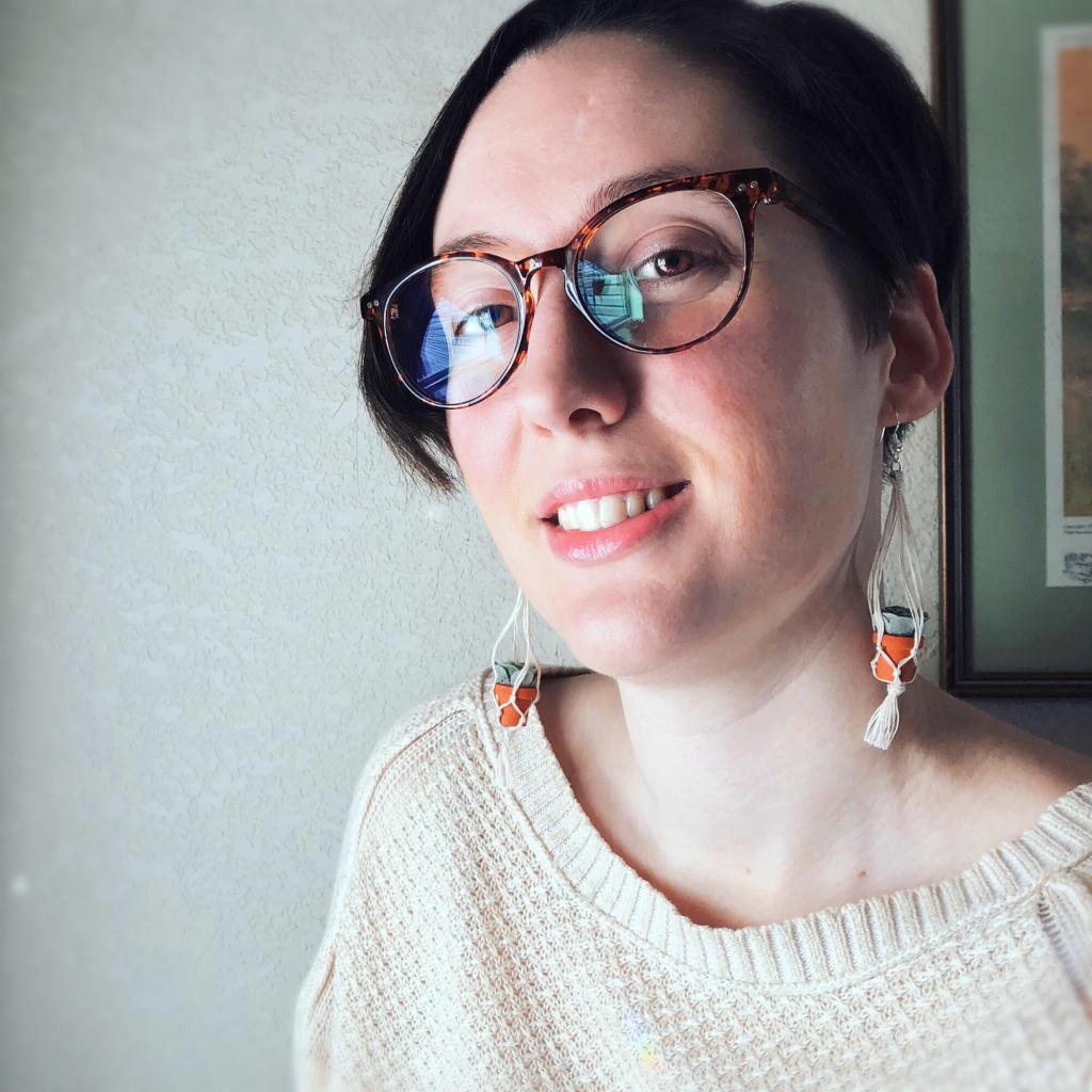 Image is a photo of Jules Arbeaux, who has glasses, pale skin, short dark hair, and dangly earrings of succulents with mini macrame hangers around them
