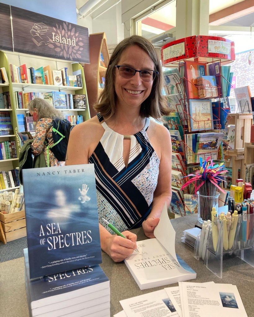 Image is a photo of author Nancy Taber signing copies of her book A Sea of Spectres in a bookstore, smiling